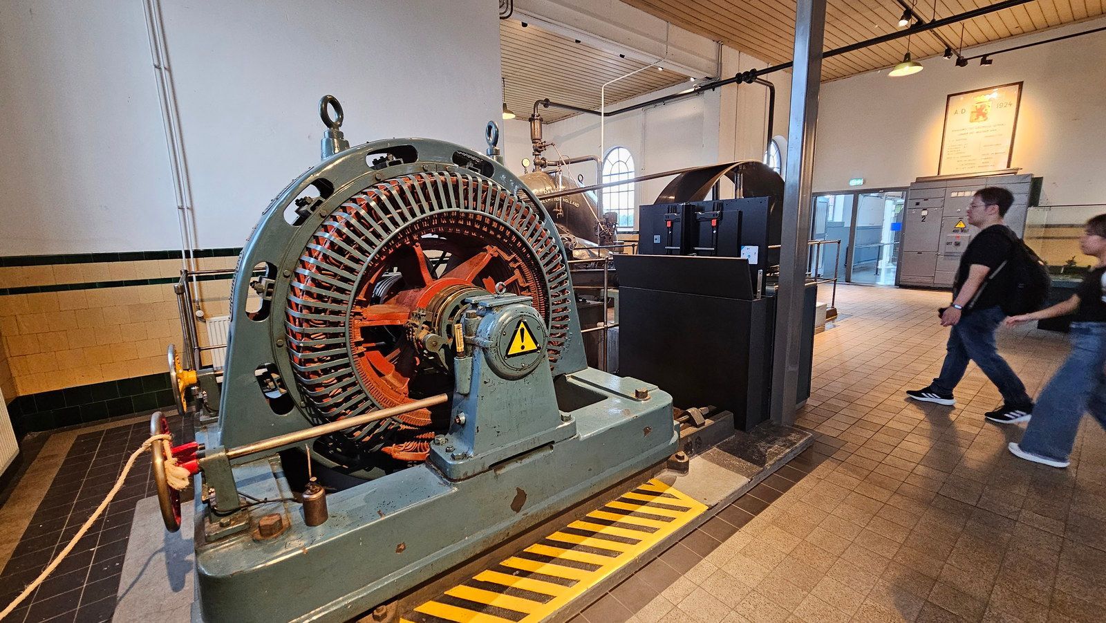 גנרטור חשמלי גדול במוזיאון, אנשים עוברים לידו. גוונים של מתכת, ירוק, כתום ואדום.