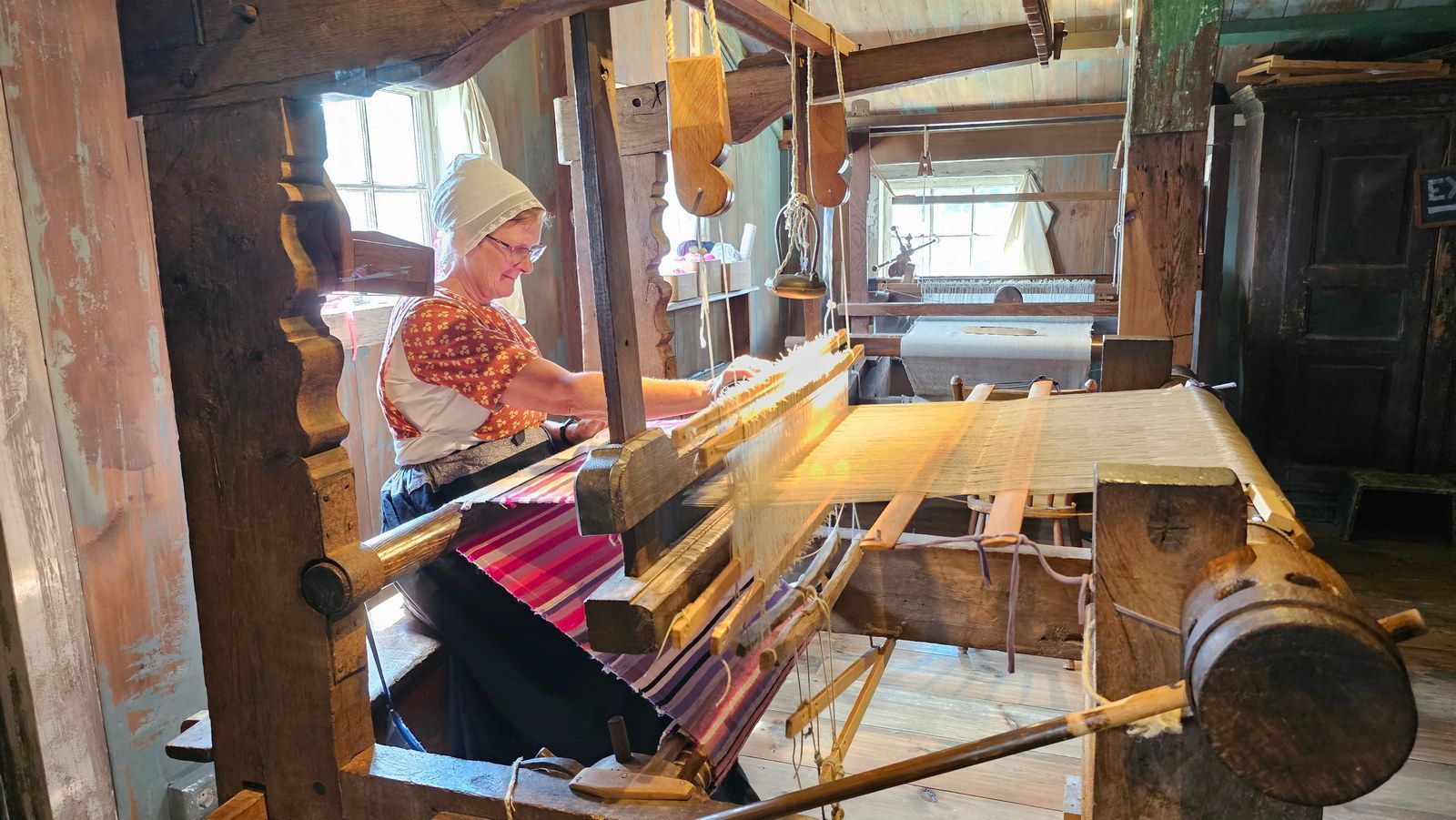 אישה אורגת על נול עץ בסדנה כפרית, עונדת כיסוי ראש ולבוש מסורתי.