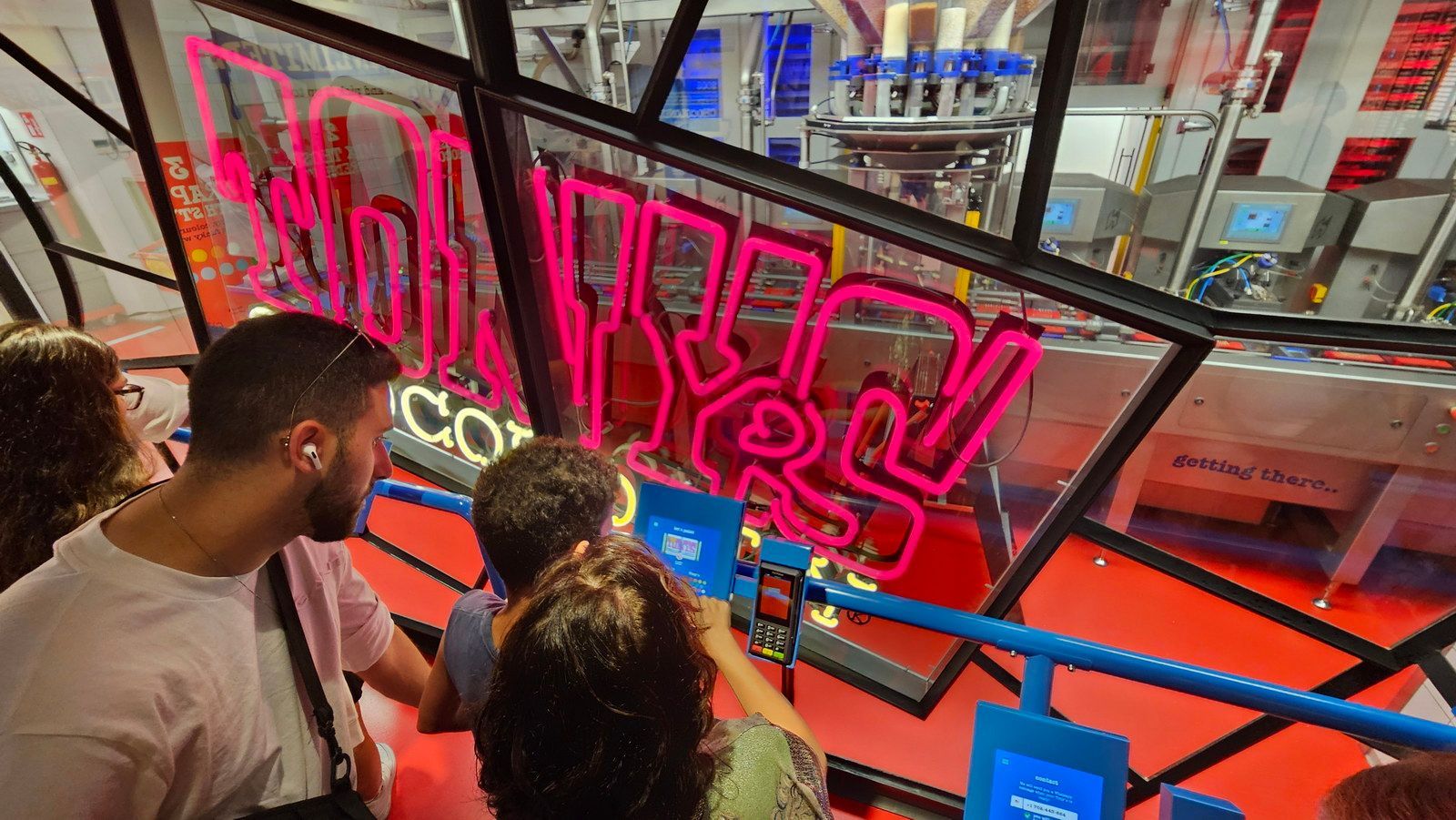 אנשים צופים במכונה של מפעל ממתקים מאחורי שלט ורוד ניאון.