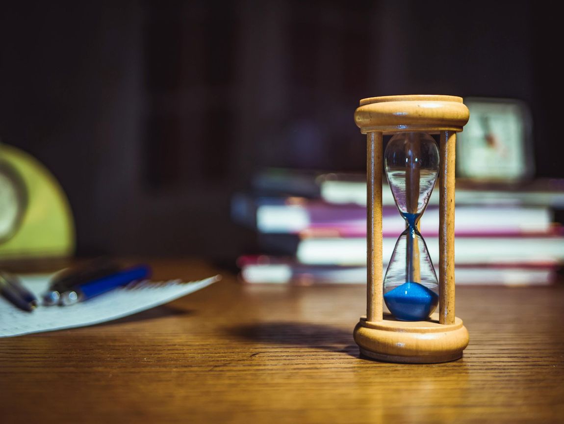 An hourglass is sitting on a wooden table in front of a stack of books.