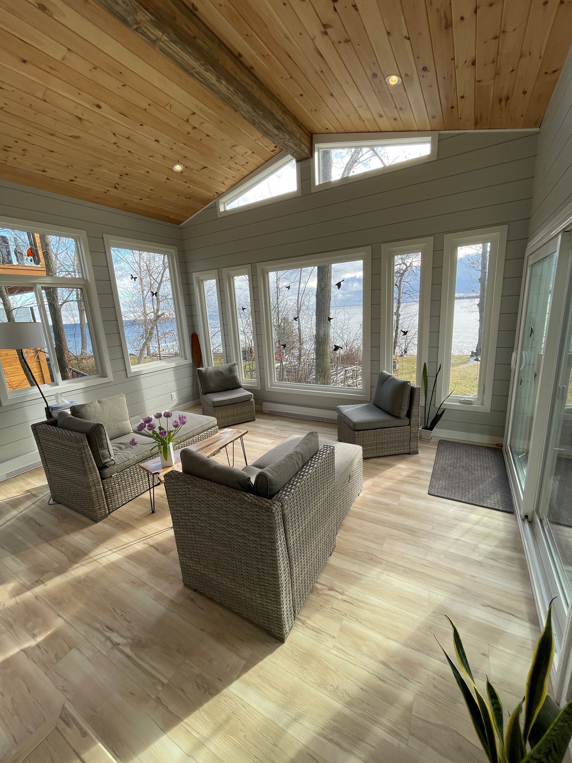 Sunroom with light wood floors, gray wicker furniture, and large windows overlooking water.