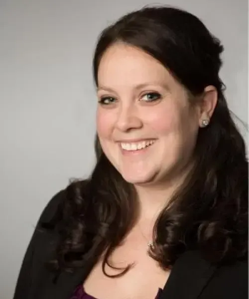 Smiling woman with dark hair and a black blazer, against a light gray background.