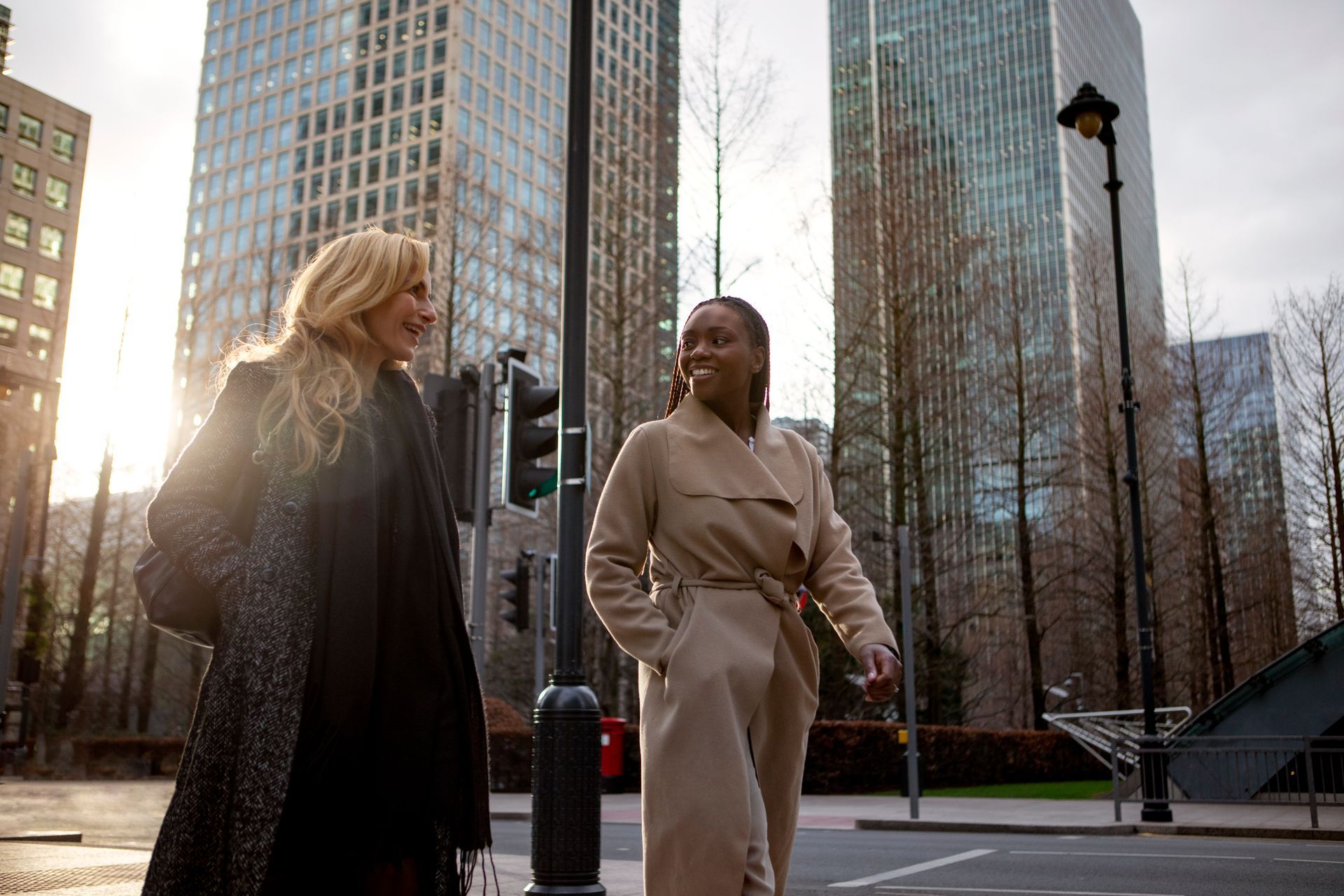 Two women walking and talking on a city street with tall buildings in the background. One woman wears a black coat; the other, a beige trench coat.