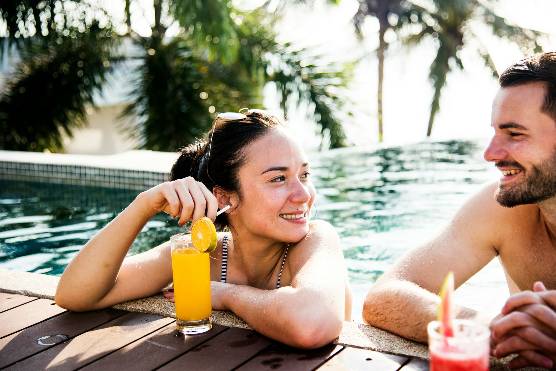 Woman and man in a pool, each with a drink, smiling and enjoying the sunshine.
