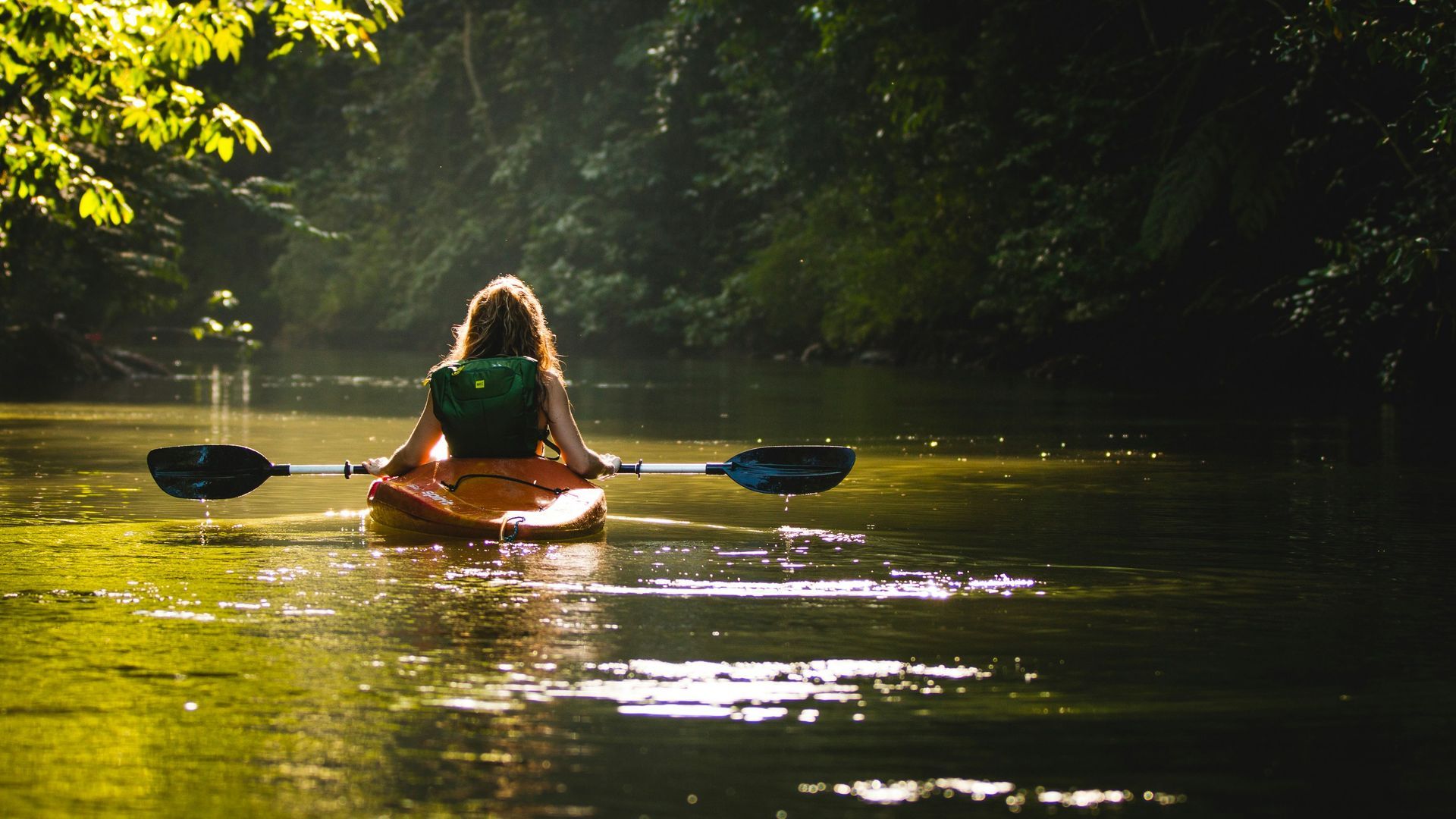 Woman kayaking on a calm river, surrounded by lush green foliage. She's paddling with two oars, wearing a green backpack.