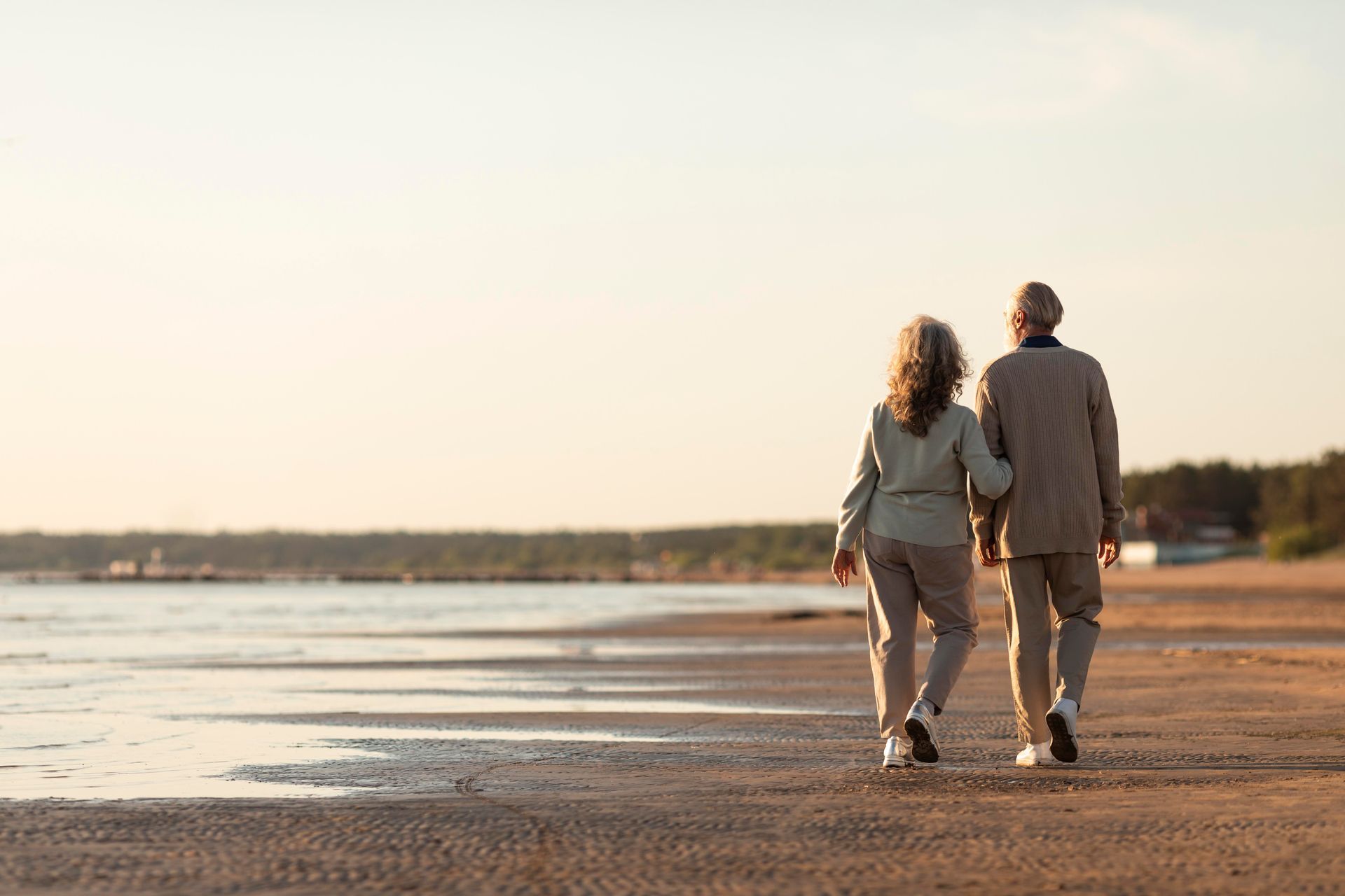 Elderly couple walks along a beach at sunset, arm in arm, silhouetted against the sky.