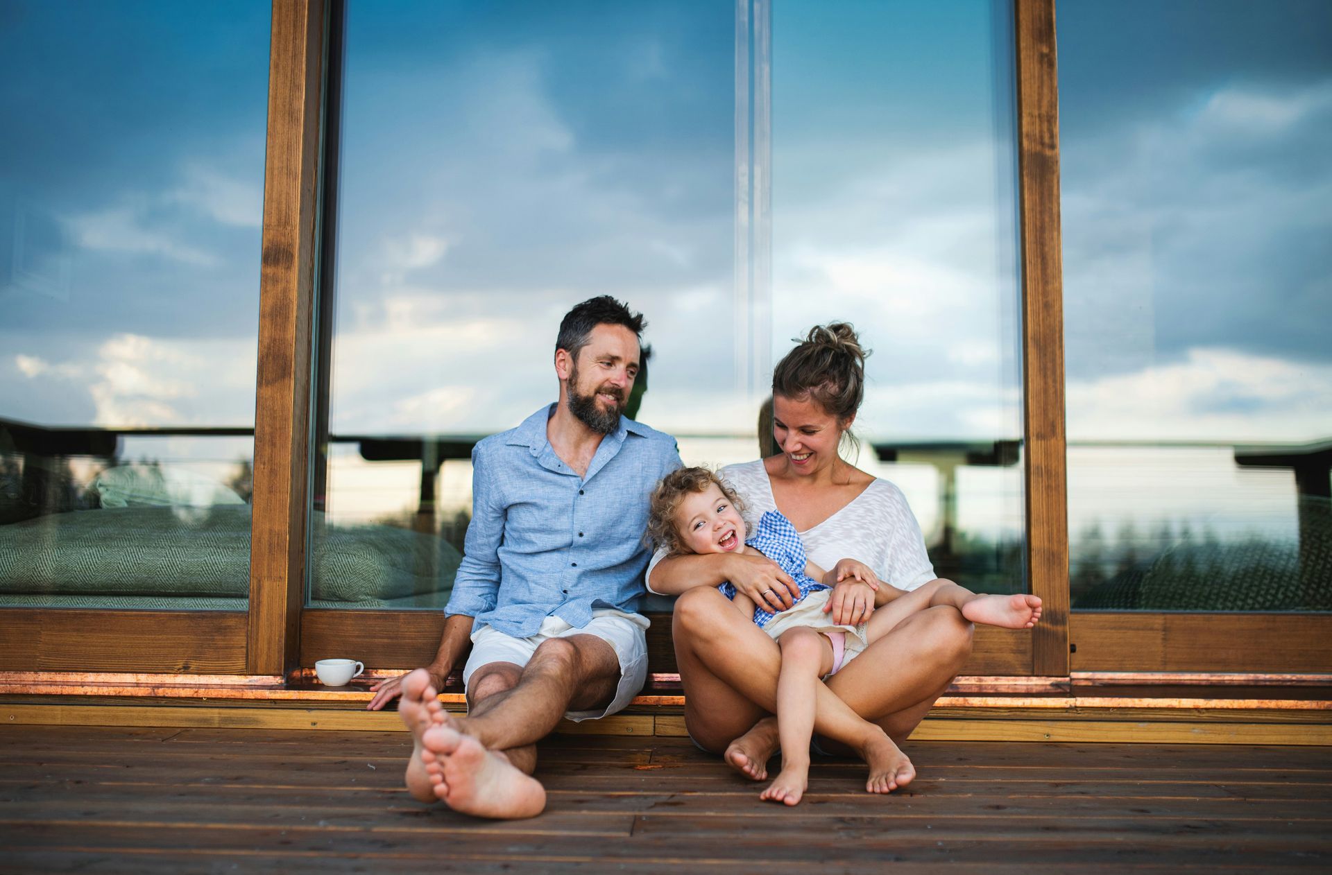 A family of three sits on a wooden deck by a large window. They are smiling and looking toward each other, with a cloudy sky in the background.
