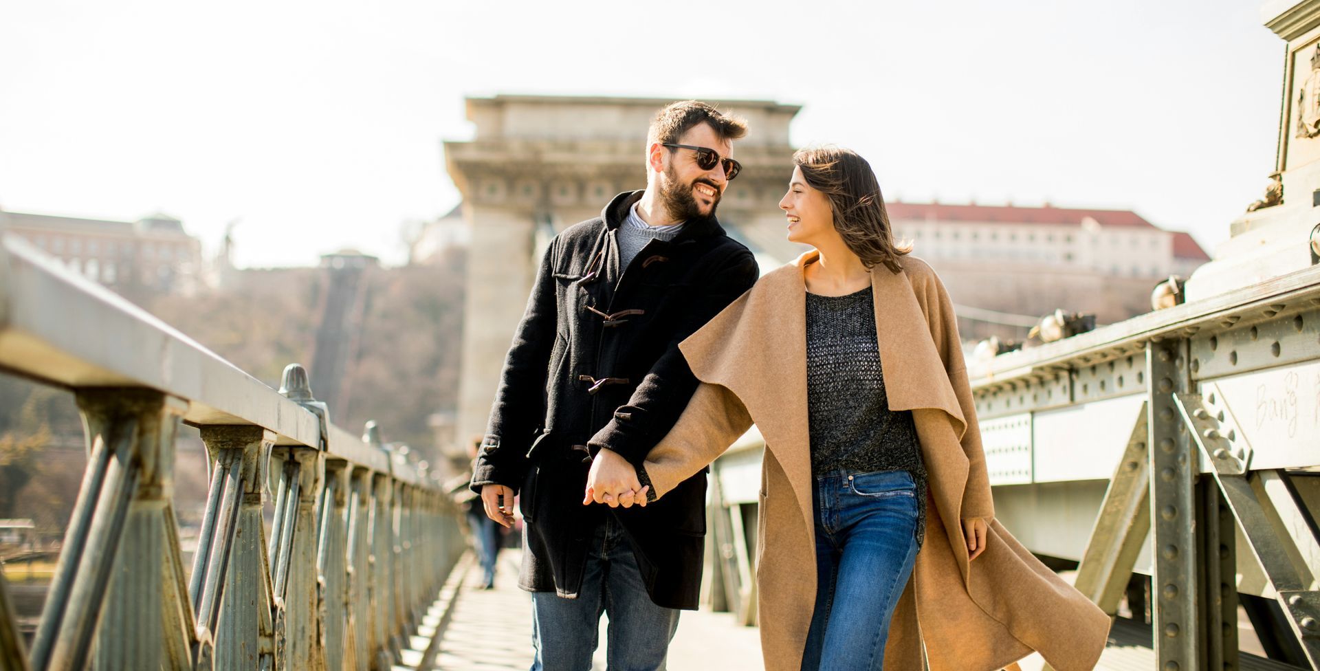 A smiling couple holds hands, walking on a bridge in the sunlight. The woman wears a tan coat, and the man wears a black coat and sunglasses.