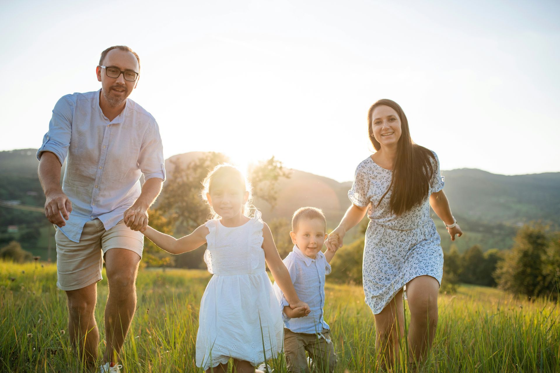 Family of four running through a grassy field toward the camera on a sunny day. Parents hold hands with two young children.