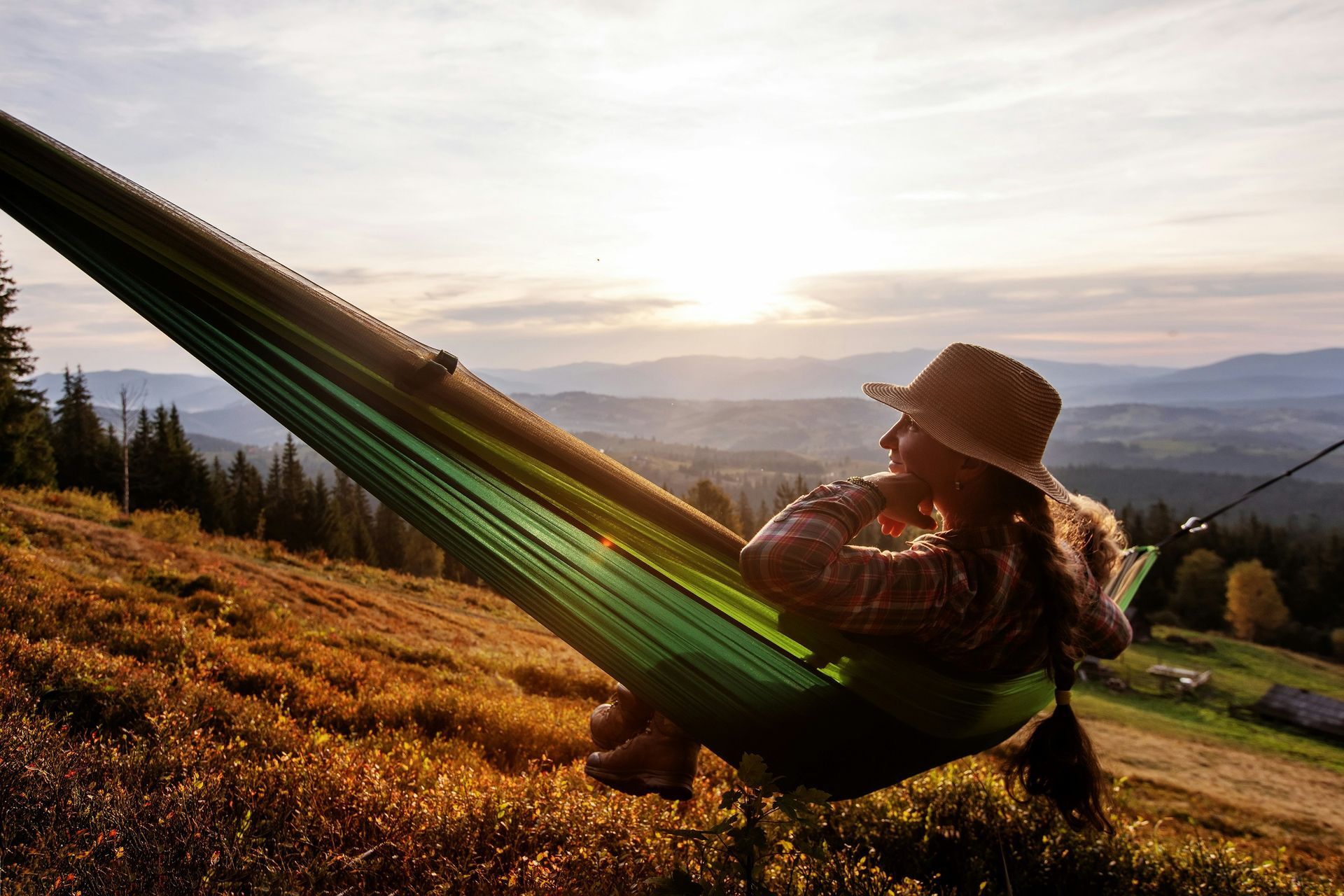 Woman in a hat relaxing in a green hammock, enjoying a scenic view of mountains and sunset.