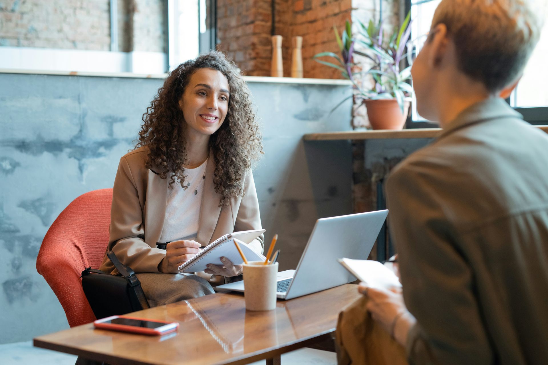 Woman with curly hair smiles, taking notes in a notebook, interviewing another person in a cafe setting.