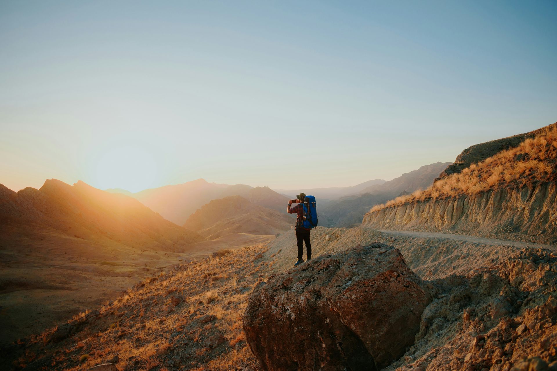A person with a backpack stands on a rocky outcrop, looking at a valley at sunset.