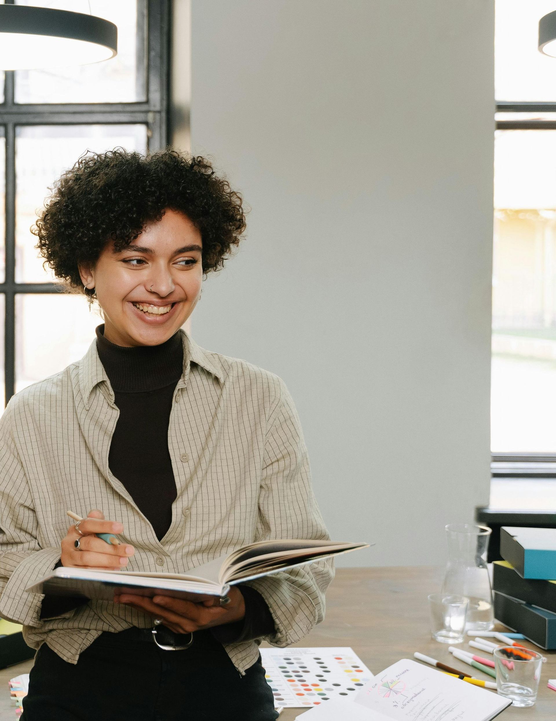 Woman with curly hair smiles, holding a notebook, in a well-lit office. She's wearing a button-down shirt over a black turtleneck.
