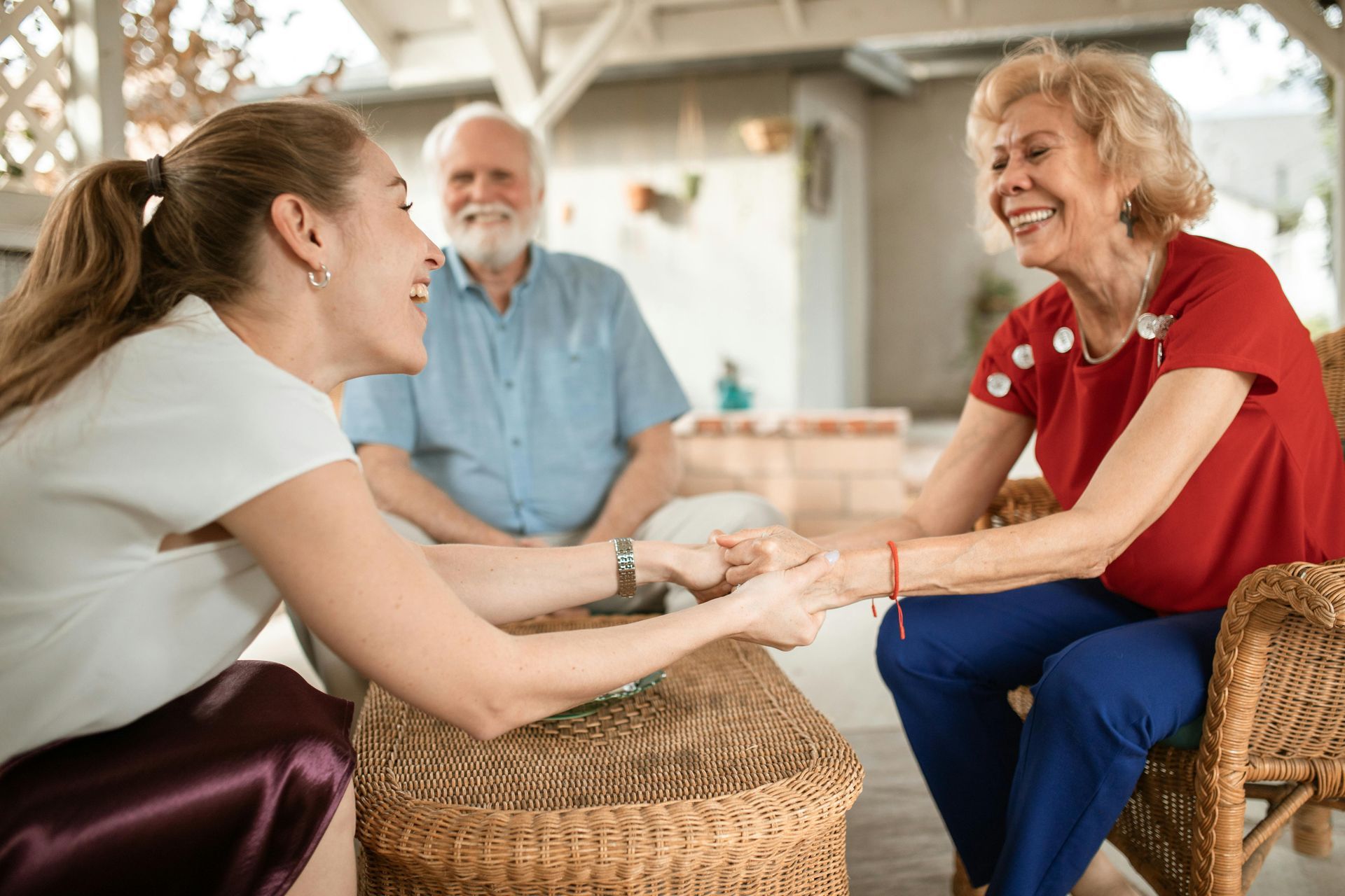 A young woman holds hands with an elderly woman, both smiling, while an elderly man smiles in the background on a patio.
