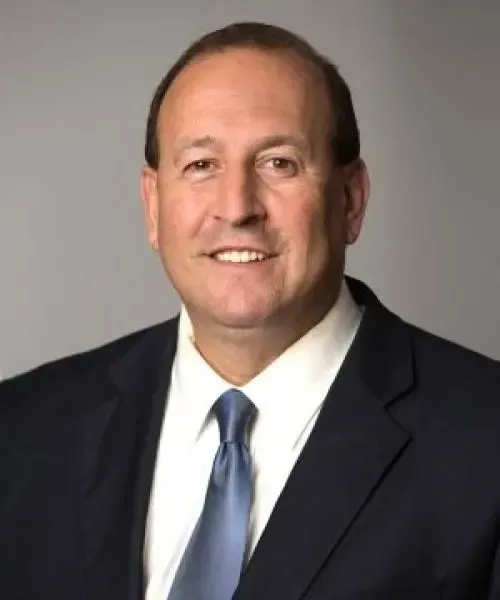 A middle-aged man in a dark suit smiles at the camera against a gray background.