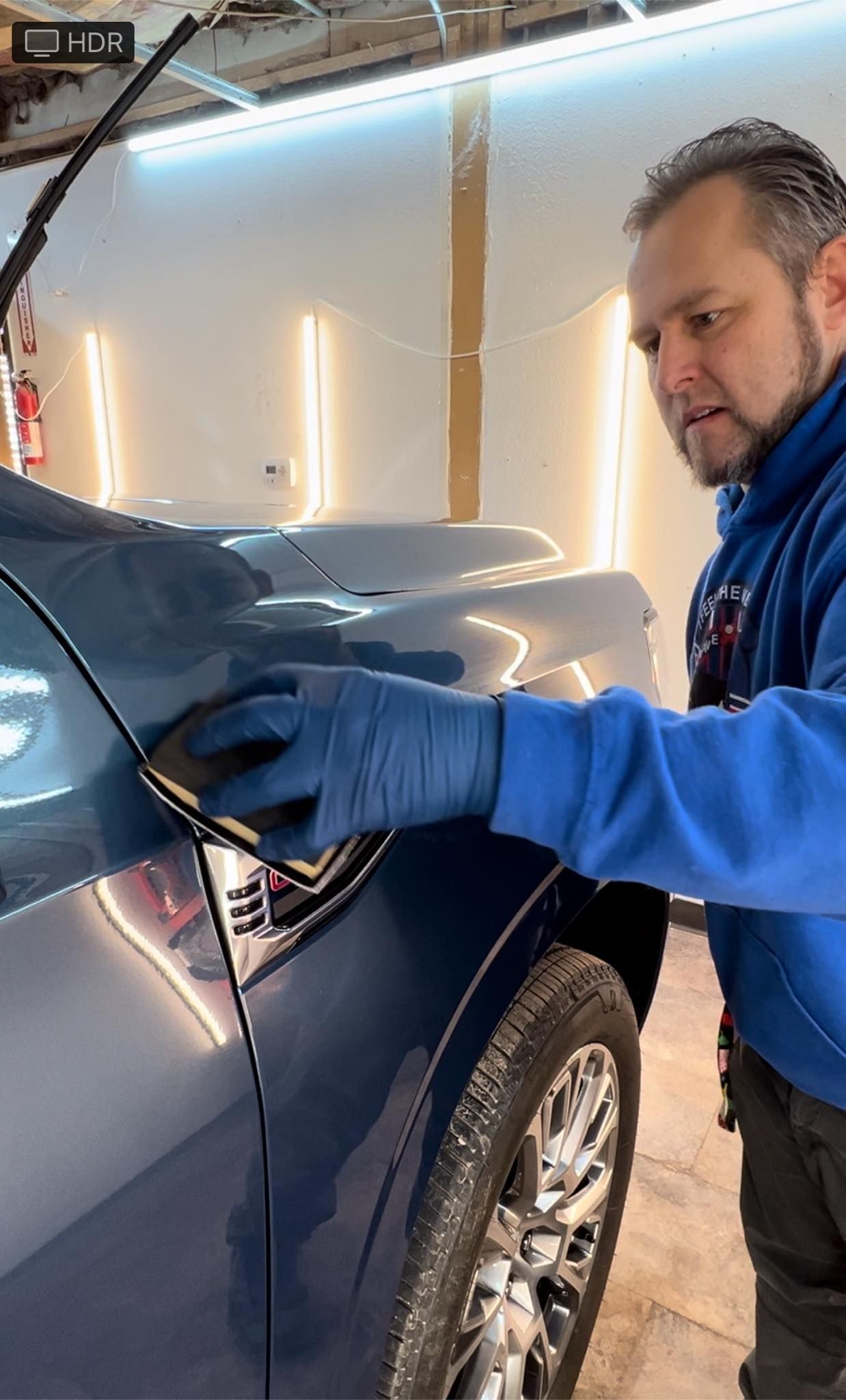 Man in blue gloves applying a product to a blue car's side panel in a well-lit garage.