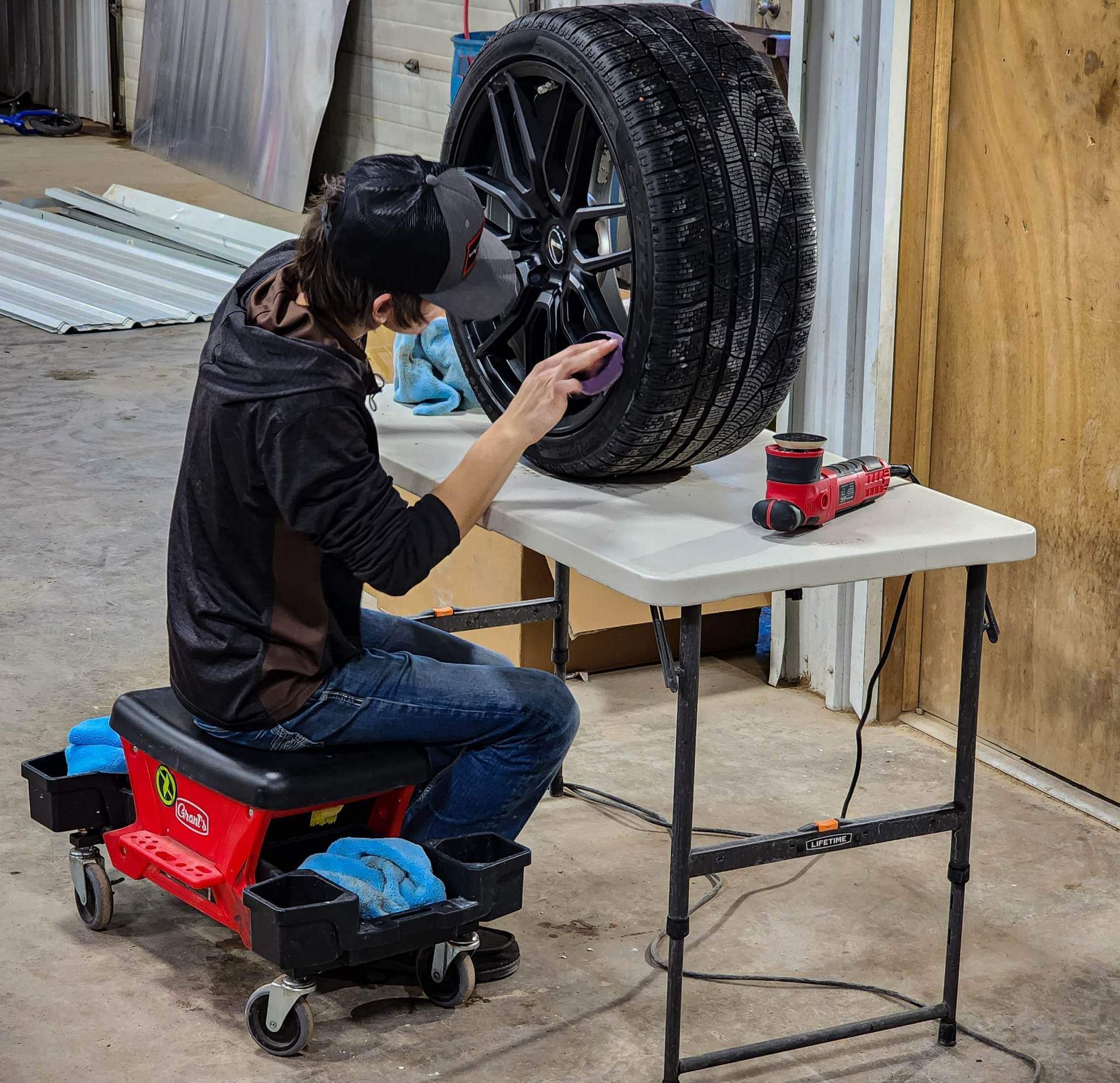 Person polishing a black car wheel on a table, seated on a rolling stool in a garage.