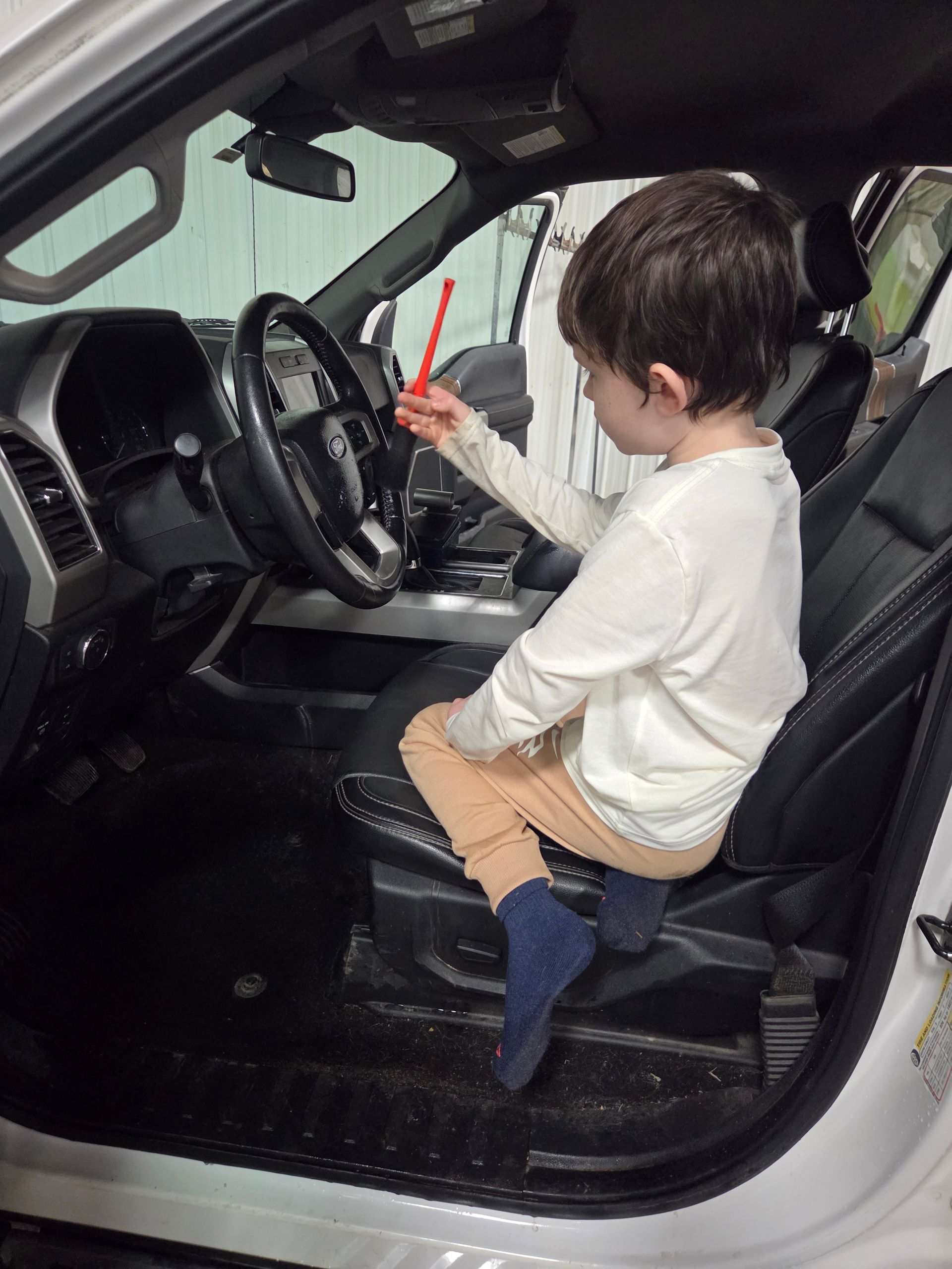 Child in a car, sitting in the driver's seat and holding a red stick.