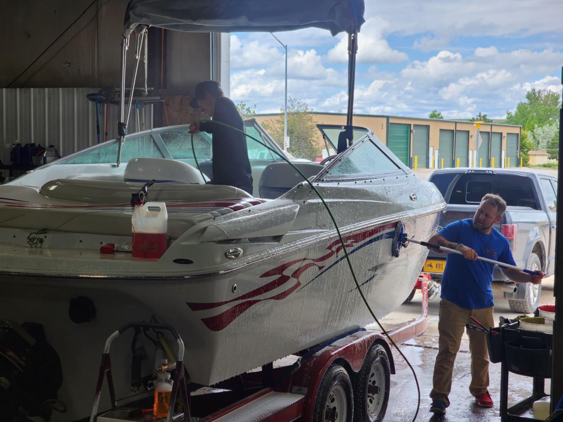 Two people washing a white boat on a trailer under a covered area. One person sprays the boat with a hose.