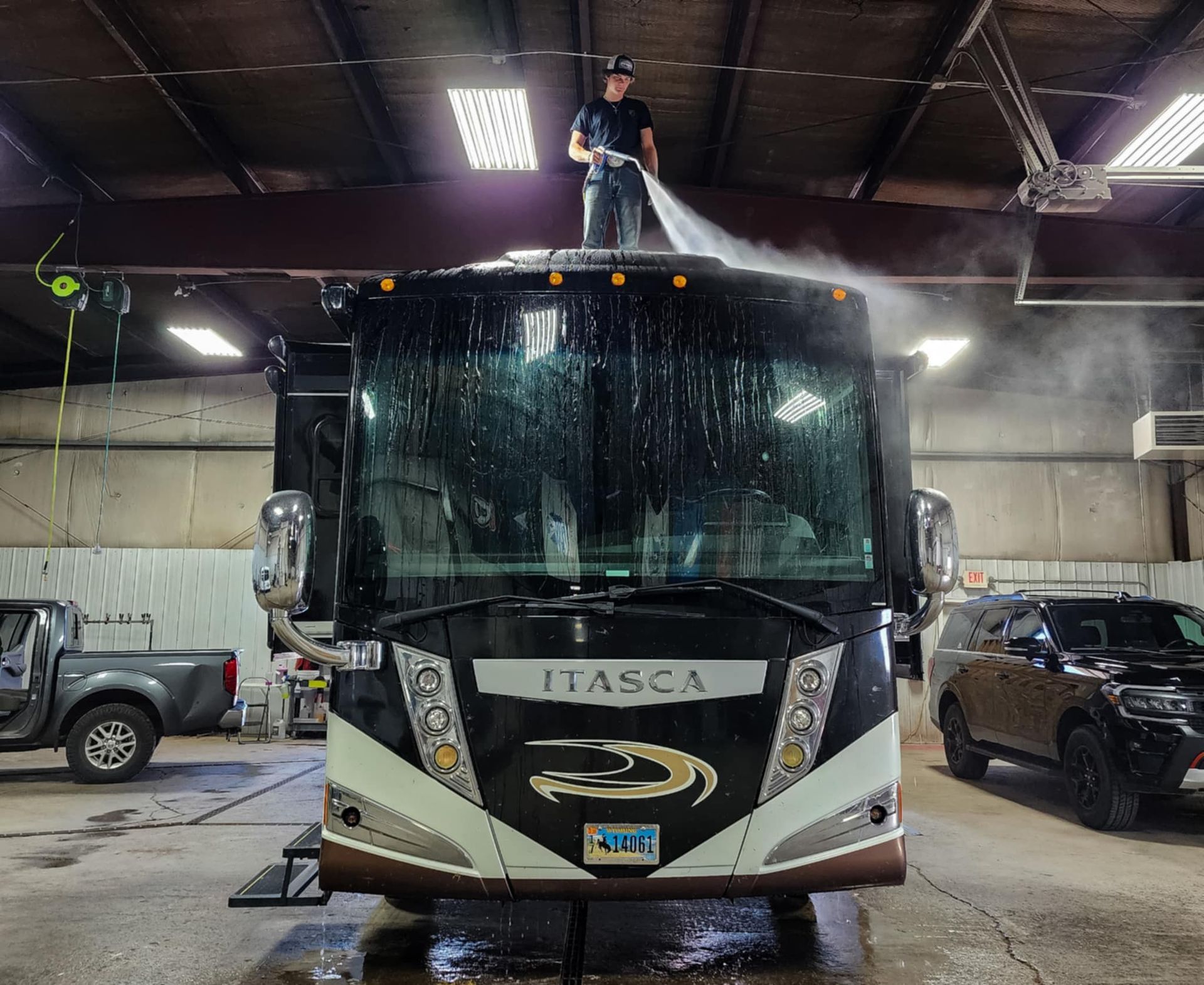 Man power washing the roof of a large RV inside a garage. The RV is black and beige with “ITASCA” on the front.