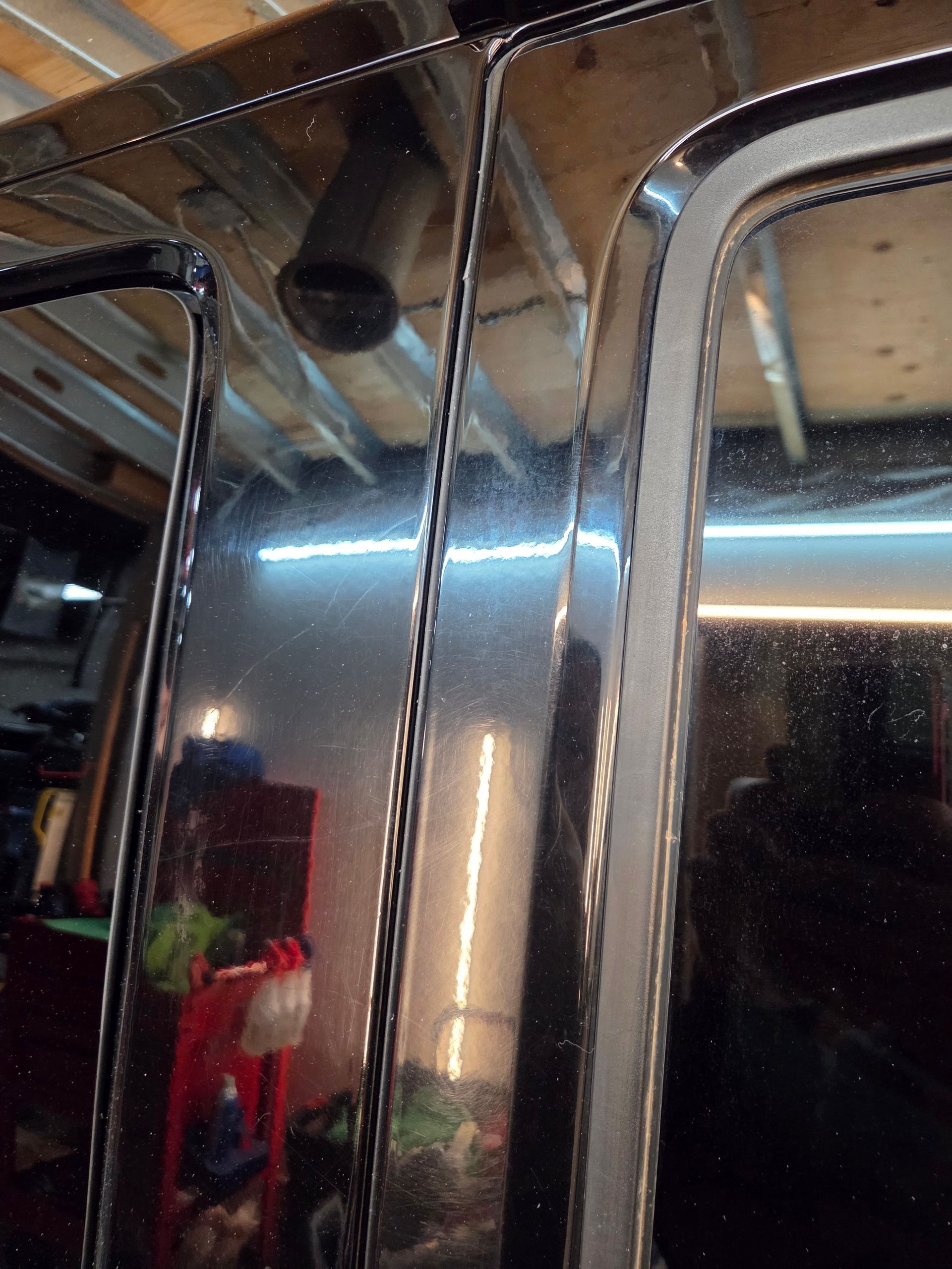 Close-up of a black vehicle's pillar, chrome trim, and side window. Workshop setting with visible tools and equipment.