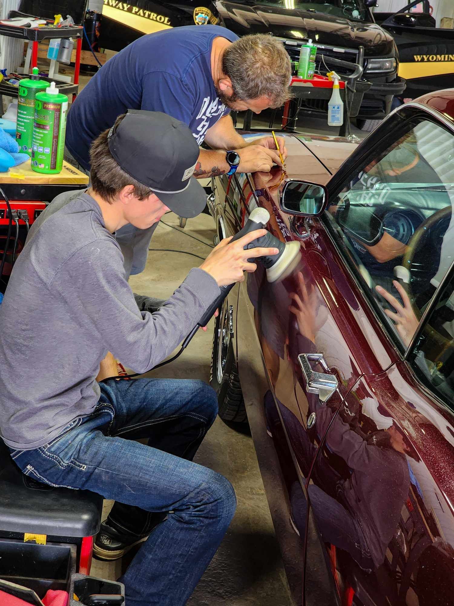 Two people polishing a maroon car in a garage. One uses a buffer, other works on trim.