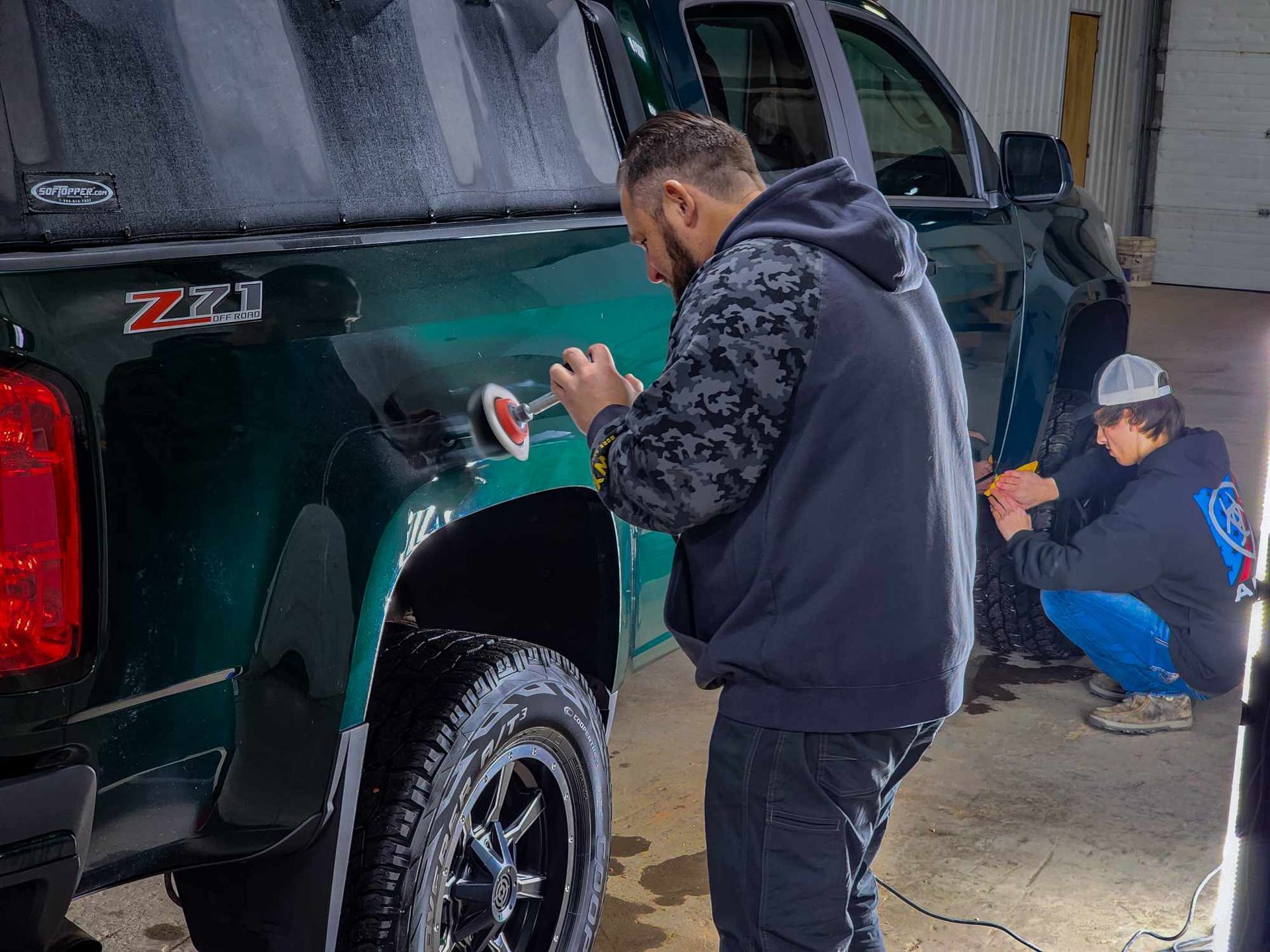 Two men working on a green truck inside a garage. One polishes the side, another works on the door.