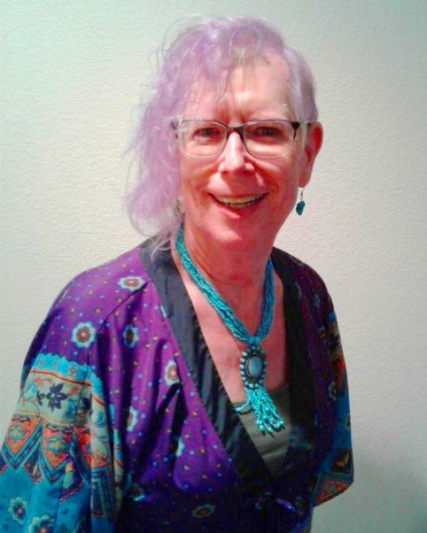 Arica King - lavender-colored hair and glasses, wearing a patterned purple top and a turquoise beaded necklace.