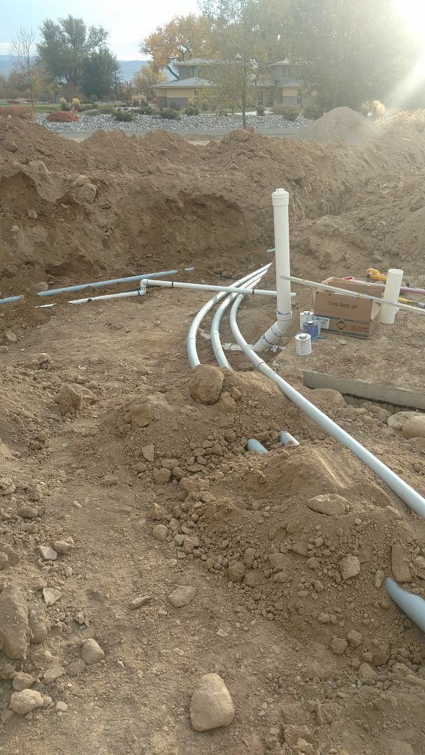 Several gray PVC pipes curve through a trench at a construction site with dirt mounds in the background.