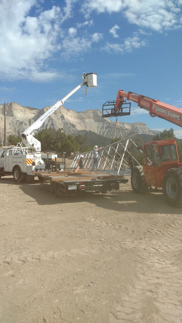 A white bucket truck and an orange telehandler lifting a metal structure in a sunny, mountainous outdoor area.