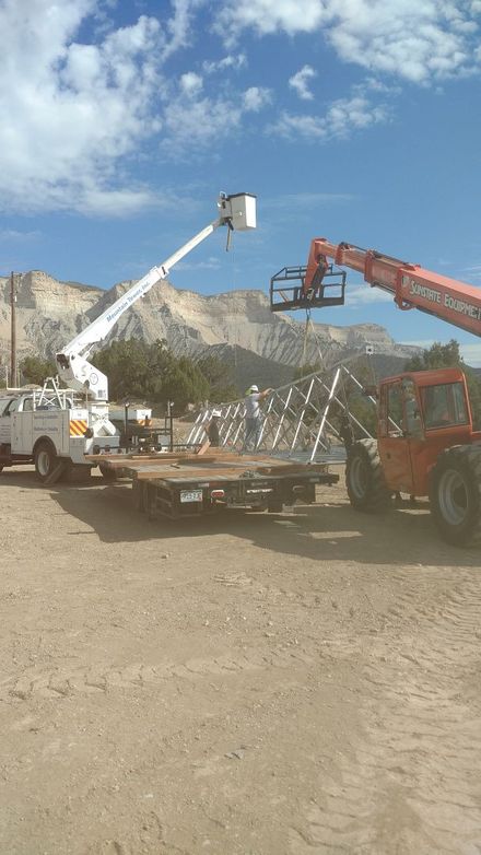 A white utility truck with an extended bucket lift and a red telehandler work on a flatbed trailer against a mountain.