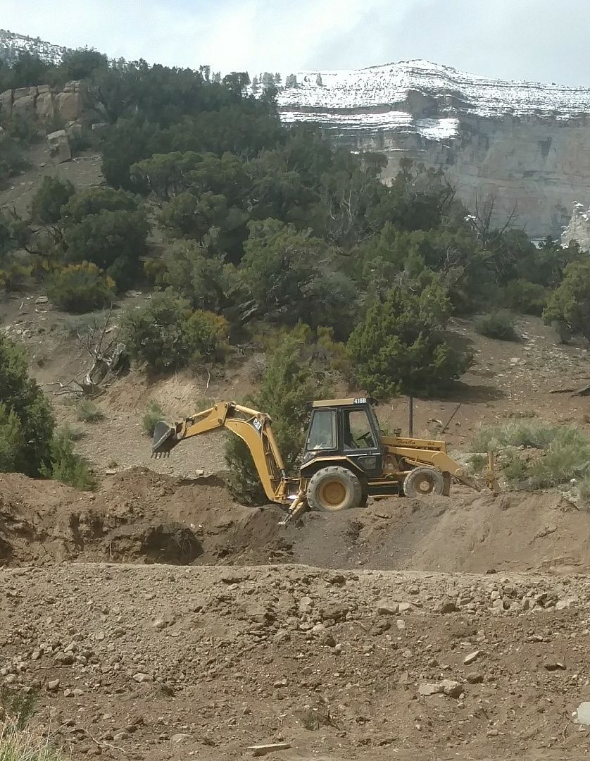 A yellow backhoe excavator works in a rocky, mountainous landscape with snow-capped peaks in the background.