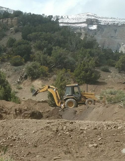 A yellow backhoe operates on a dirt slope in a mountainous, partially forested area with snow-capped peaks in the distance.
