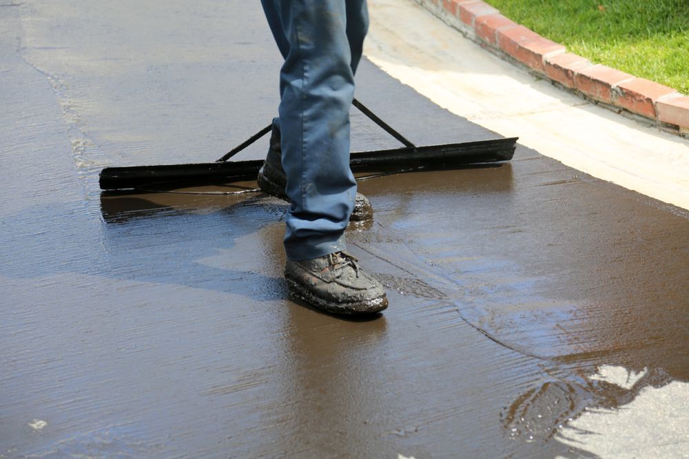 A person uses a squeegee to spread black asphalt sealant across a driveway.