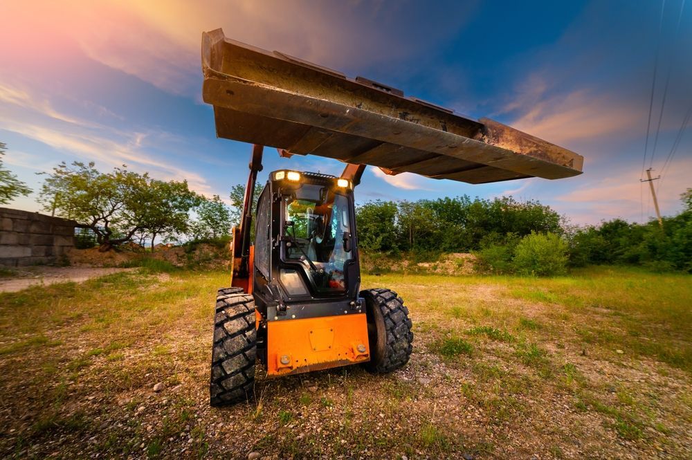 Orange skid steer loader with a raised bucket in a rural, grassy field during a sunset.