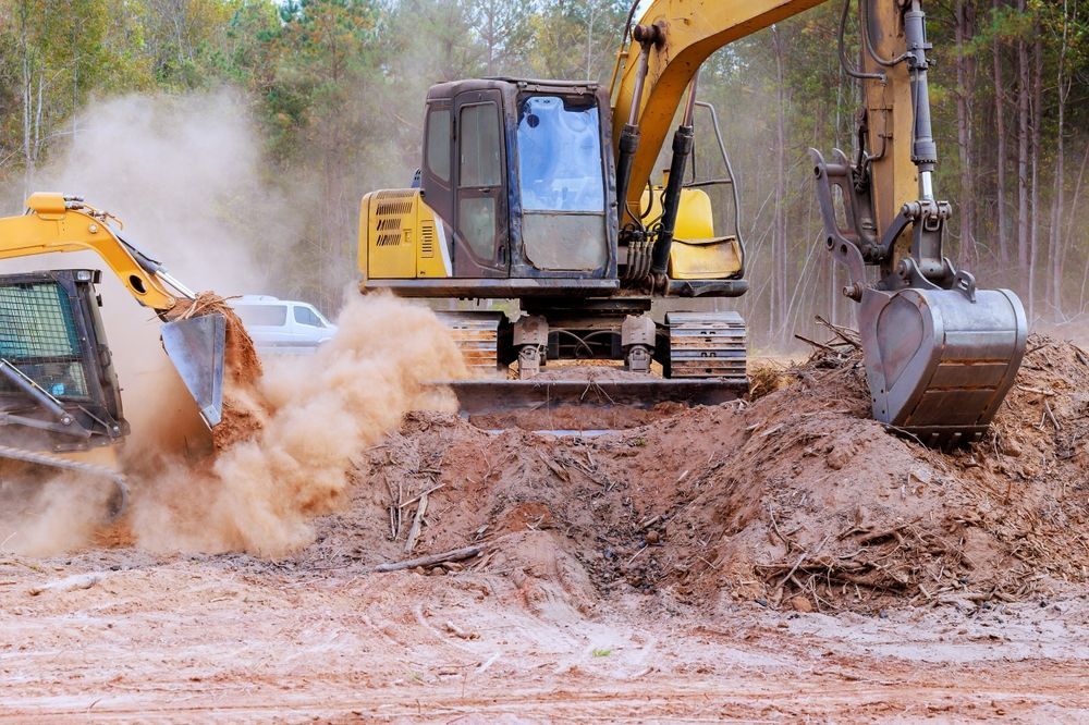 Two yellow excavators stir up a cloud of dust while digging in a wooded area.