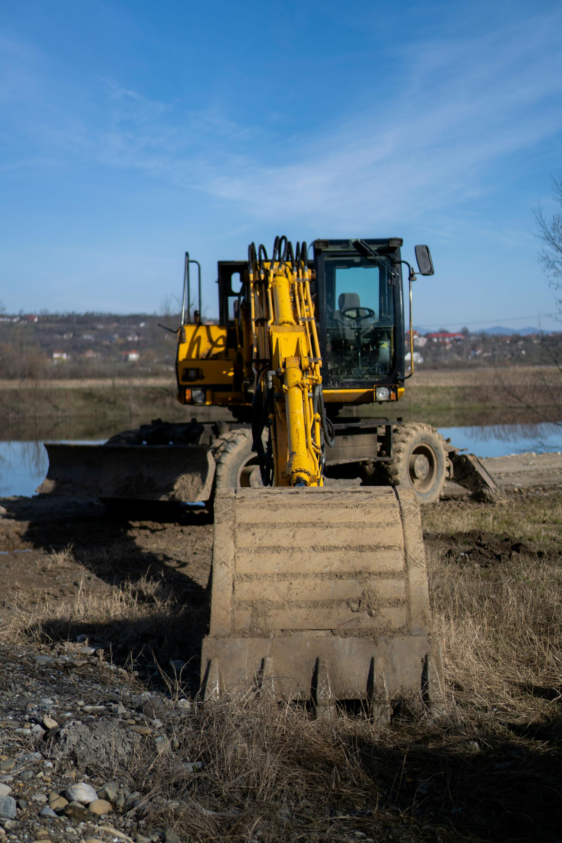 A yellow wheeled excavator parked on dirt near water under a clear blue sky.