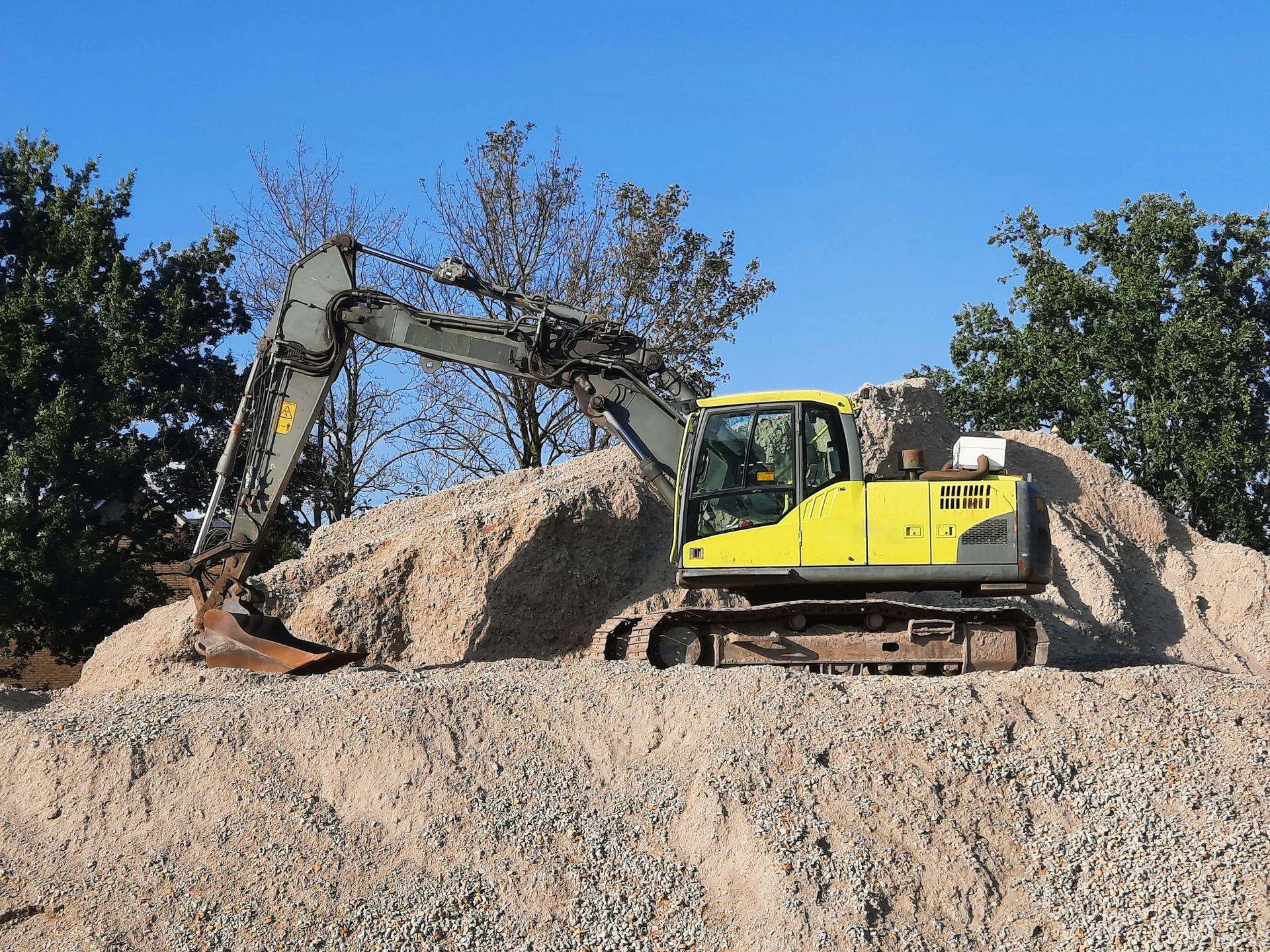 A yellow excavator with its boom extended, parked atop a large pile of gravel against a clear blue sky.