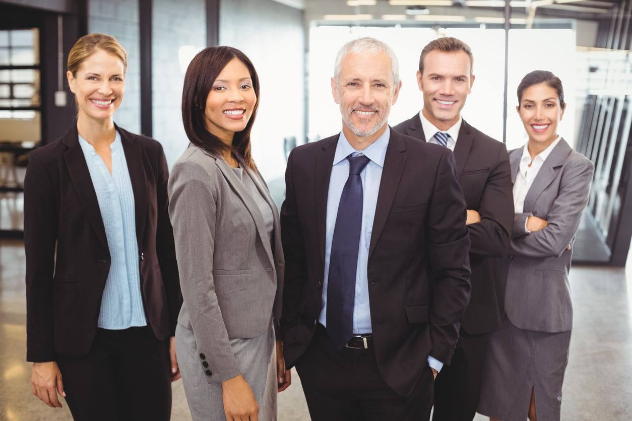 Group of five professionals, diverse in gender and age, smiling and looking at the camera, office setting.