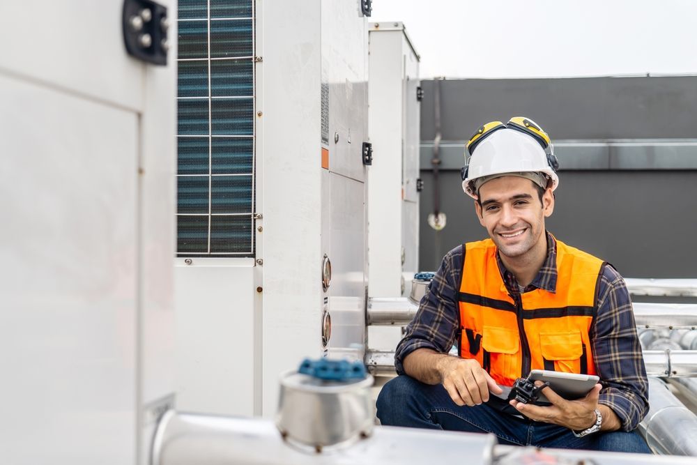A man in a hard hat and orange vest is sitting on a pipe holding a tablet.