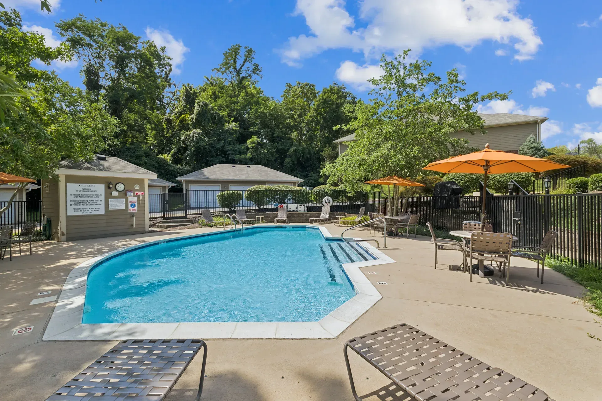 Swimming pool with lounge chairs, orange umbrellas, and small buildings; trees in background.