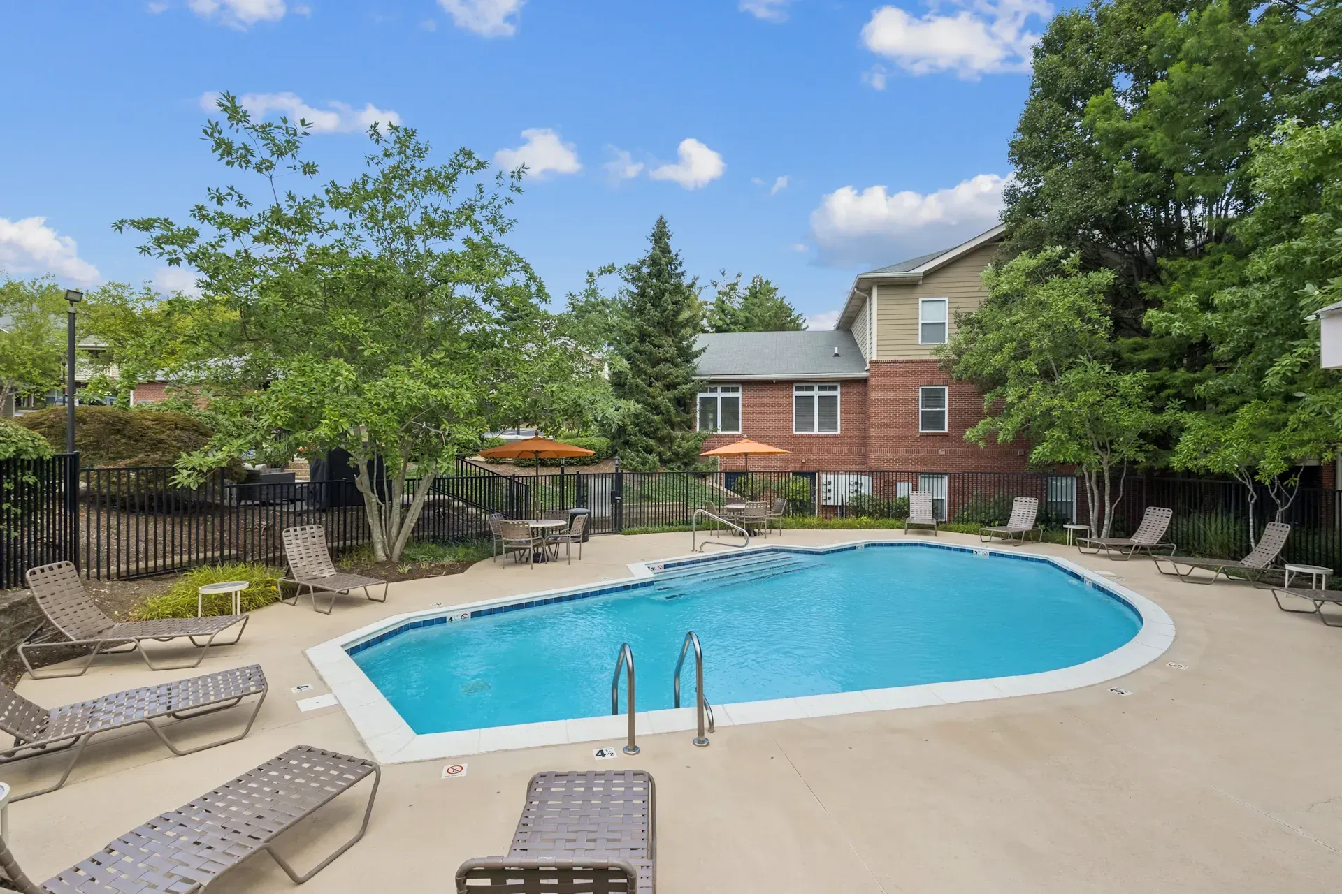 Swimming pool with lounge chairs, surrounded by trees and a brick building, under a blue sky.