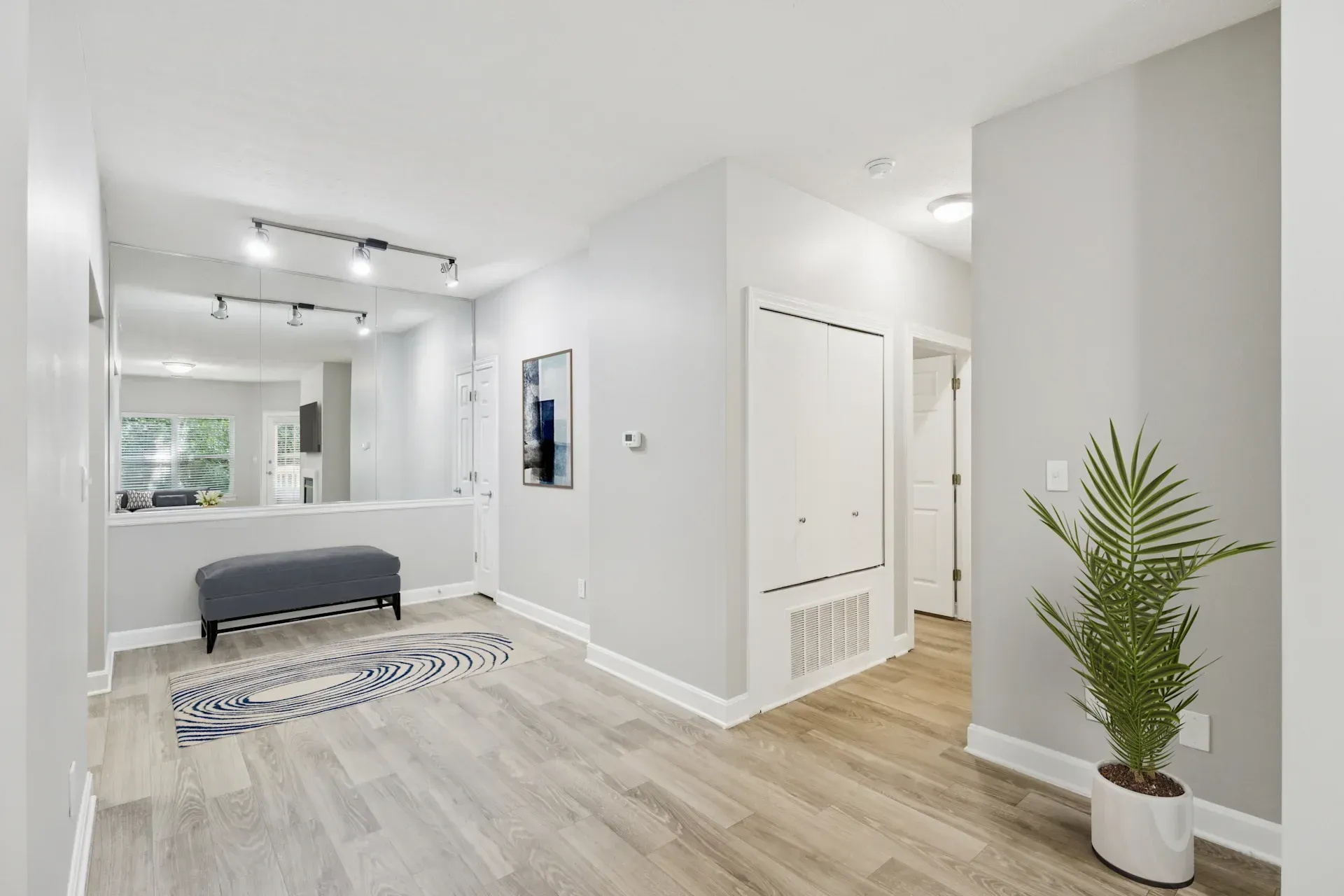 Bright hallway with light gray walls, wood-look flooring, and a large mirror. A bench and plant are visible.