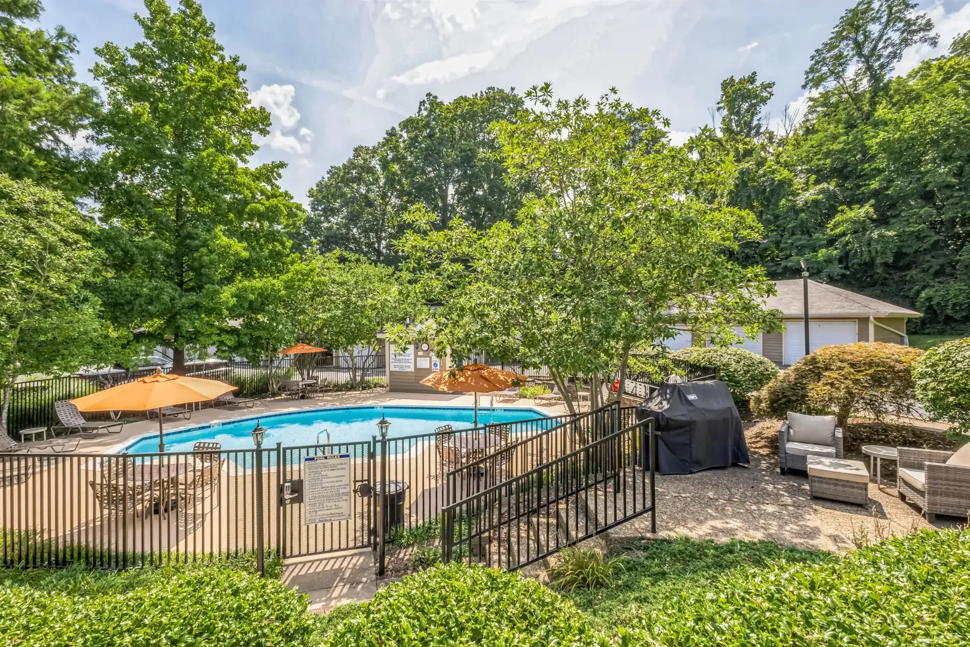 Pool area with fence, lounge chairs, umbrellas, and trees. Grill and building visible.
