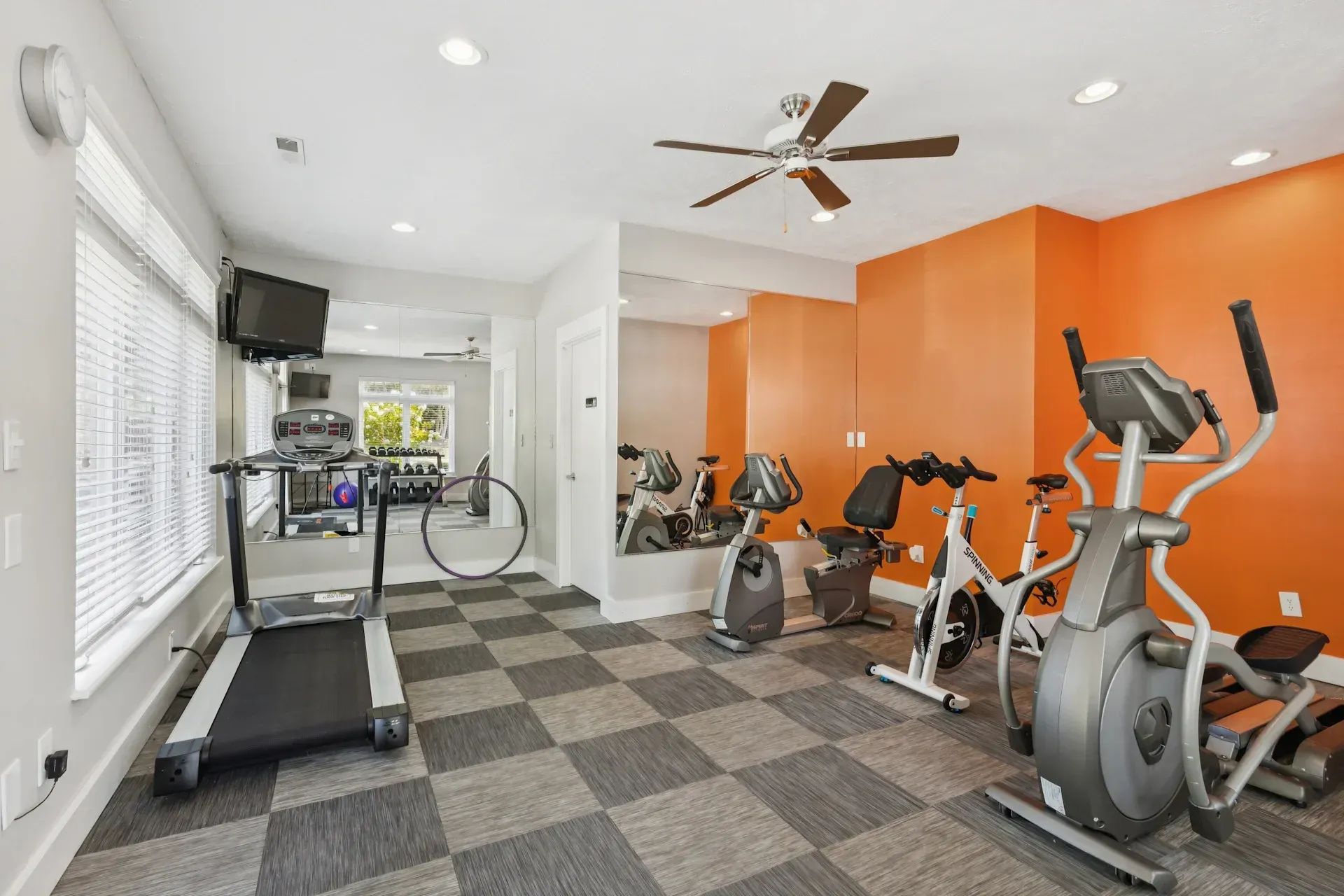 Home gym with cardio machines: treadmill, elliptical, and stationary bikes. Orange wall, checkered floor, large mirror.