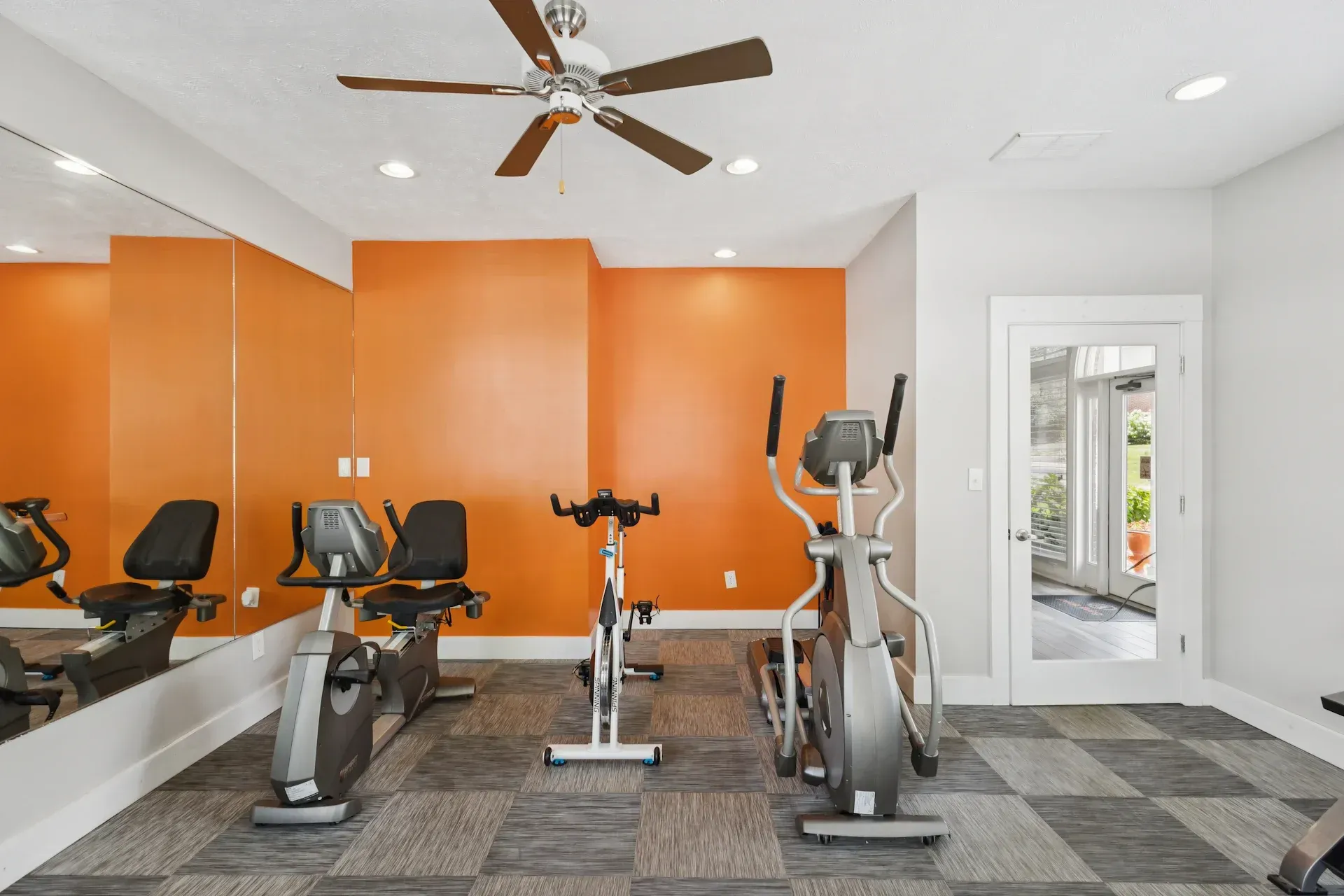 Gym with exercise bikes and elliptical machine. Orange accent wall, mirror, and gray checkered flooring.
