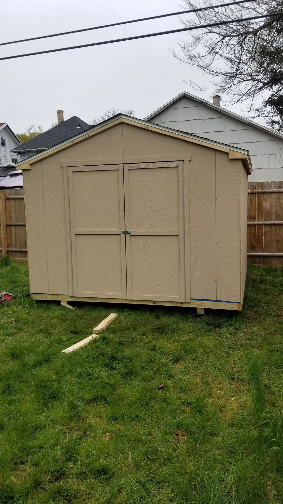Beige garden shed with double doors on a grassy backyard, near a wooden fence and house.