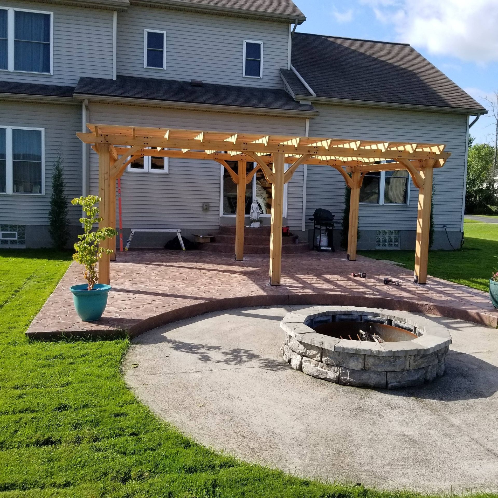 Wooden pergola over a brick patio beside a stone fire pit in a suburban backyard