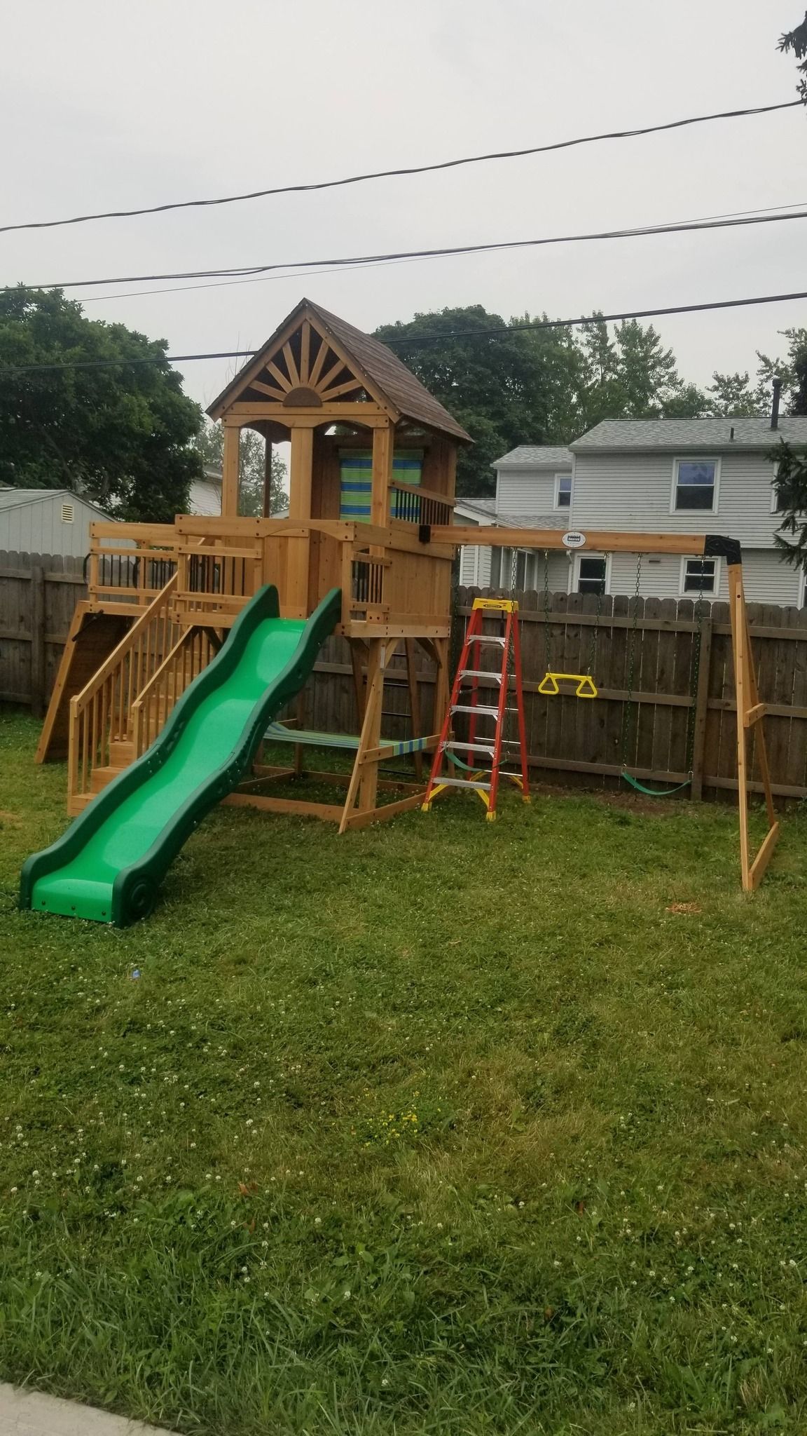 Wooden backyard playset with green slide, ladder, and small lookout under an overcast sky