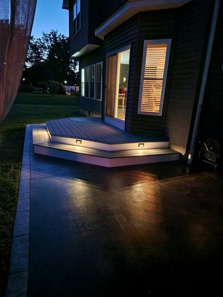 Lit wooden deck and steps beside a house at dusk, with warm indoor light reflecting on the wet driveway.